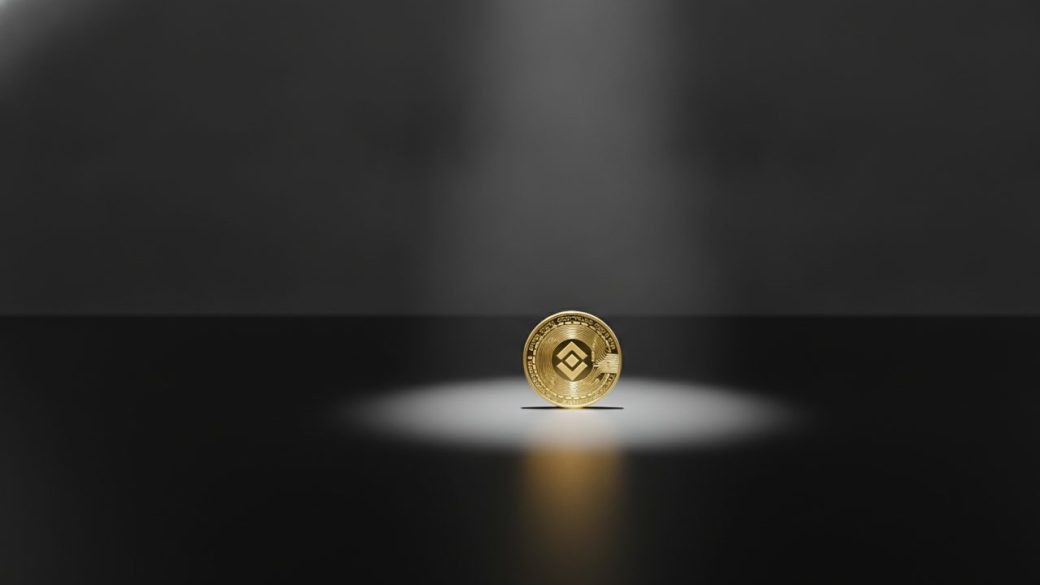 A single glowing Binance crypto coin on a dark surface under a spotlight, casting a long dramatic shadow, clean uncluttered background, high-contrast composition, no text or watermark