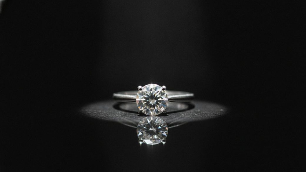 A single sparkling diamond ring on a dark reflective surface under a dramatic spotlight, sharp high-contrast lighting, clean uncluttered background, no text or logos
