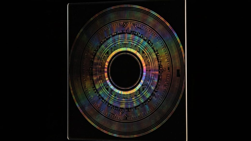 A single gleaming silicon semiconductor wafer held upright against a clean dark background, dramatic side lighting catching its rainbow circuitry, sharp high contrast, no text