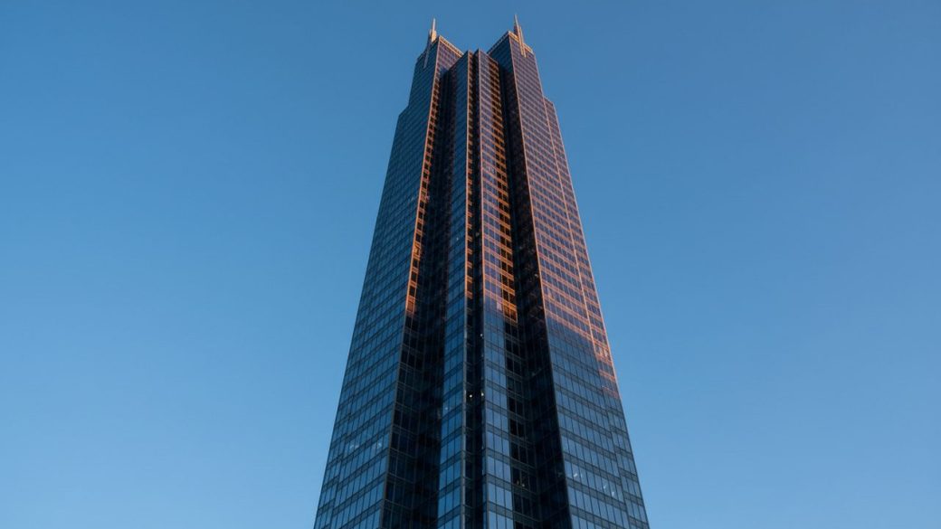 A single tall corporate glass skyscraper rising against a clear sky, dramatic low-angle lighting, high contrast, photorealistic
