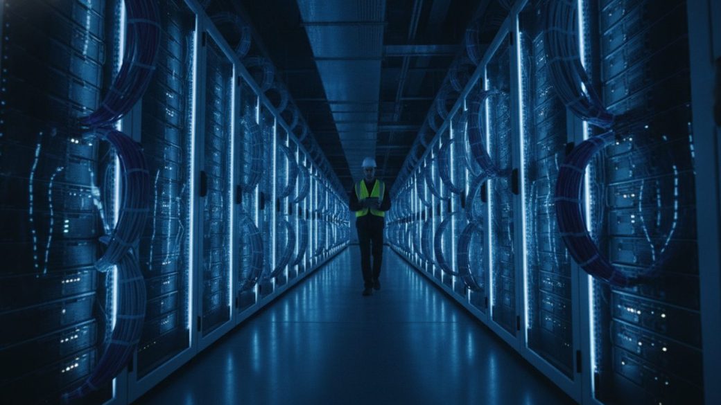 A vast modern data centre interior with long rows of glowing server racks and blue cabling stretching into the distance, one engineer in a hard hat walking the aisle