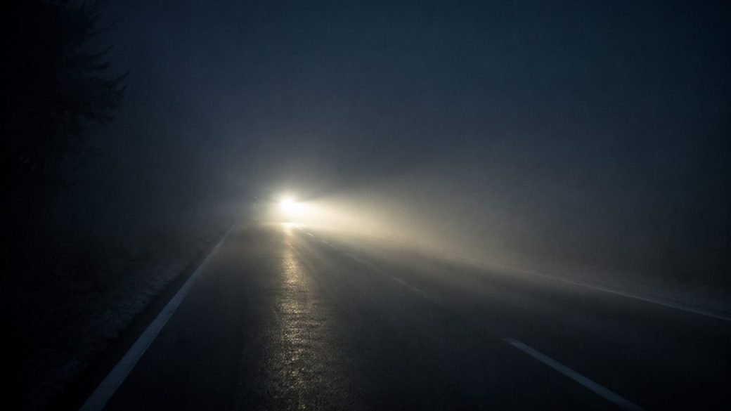 A lone car headlight cutting through heavy night fog on an empty highway, single focal light in dramatic high contrast.
