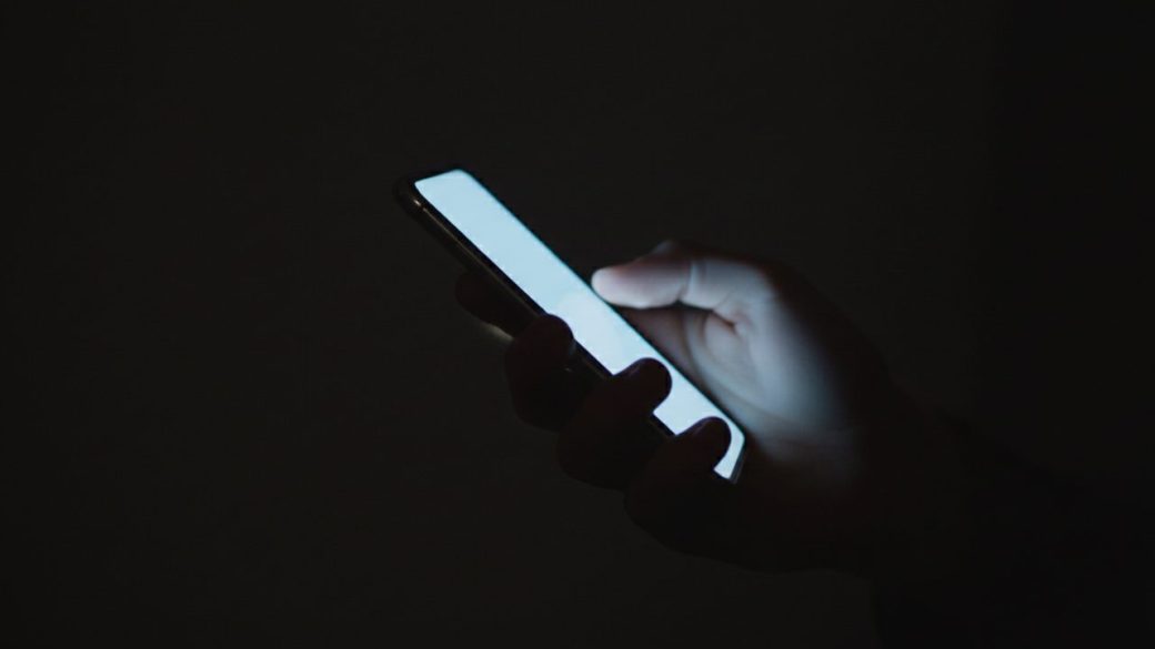 A young teenager's hand holding a glowing smartphone in a dark room, single focal subject, dramatic high-contrast lighting, clean background